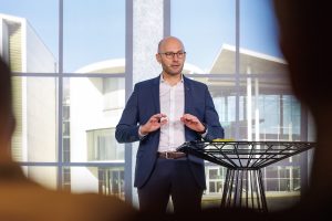 Ein Mann im blauen Anzug und weißem Hemd steht auf einer Bühne hinter einem modernen Stehtisch und spricht mit erhobenen Händen gestikulierend zu einem Publikum. Im Hintergrund sind große Fensterflächen mit Blick auf moderne Architektur zu sehen.