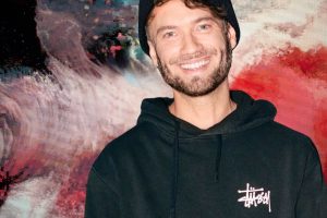 Porträt eines lächelnden Mannes mit kurzem Bart und schwarzer Strickmütze. Er trägt einen schwarzen Hoodie mit weißem Logo auf der Brust. Im Hintergrund ist ein großformatiges, abstraktes Kunstwerk in kräftigen Rot-, Weiß-, Schwarz- und Blautönen zu sehen.