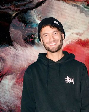 Porträt eines lächelnden Mannes mit kurzem Bart und schwarzer Strickmütze. Er trägt einen schwarzen Hoodie mit weißem Logo auf der Brust. Im Hintergrund ist ein großformatiges, abstraktes Kunstwerk in kräftigen Rot-, Weiß-, Schwarz- und Blautönen zu sehen.