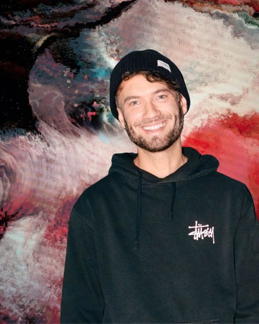 Porträt eines lächelnden Mannes mit kurzem Bart und schwarzer Strickmütze. Er trägt einen schwarzen Hoodie mit weißem Logo auf der Brust. Im Hintergrund ist ein großformatiges, abstraktes Kunstwerk in kräftigen Rot-, Weiß-, Schwarz- und Blautönen zu sehen.