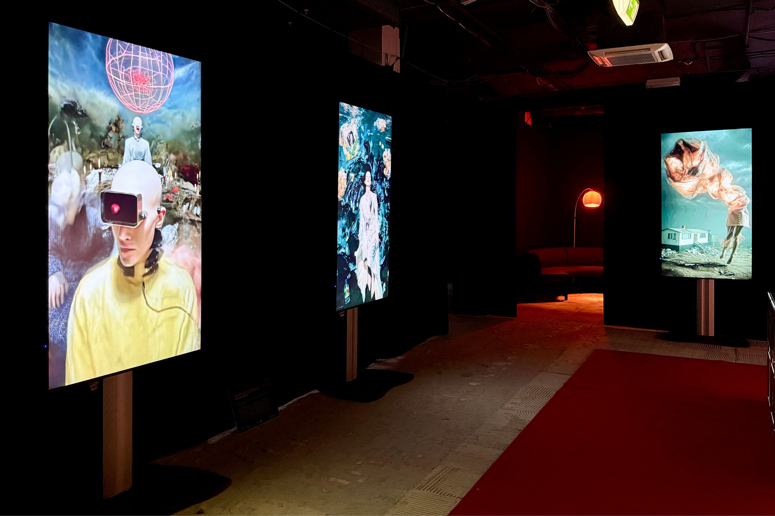 Dunkel inszenierter Ausstellungsraum mit drei hochformatigen, leuchtenden Screens auf Podesten. Die digitalen Kunstwerke zeigen surreale Motive in kräftigen Farben. Ein roter Teppich führt durch den Raum, im Hintergrund steht eine Lounge mit Sofa und roter Stehlampe.