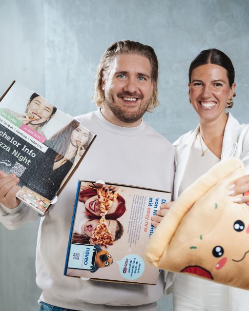 Zwei lächelnde Personen stehen vor neutralem Hintergrund und halten geöffnete Pizzakartons mit Werbeaufdruck sowie ein großes, kuscheliges Pizzastück-Kissen in die Kamera.