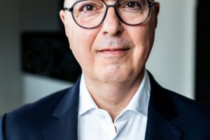 Porträt eines Mannes mit Glatze und runder, dunkler Brille. Er trägt ein weißes Hemd und ein dunkles Sakko und blickt freundlich und direkt in die Kamera. Der Hintergrund ist unscharf und wirkt wie ein modernes Büro- oder Innenraumsetting.