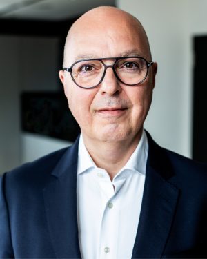 Porträt eines Mannes mit Glatze und runder, dunkler Brille. Er trägt ein weißes Hemd und ein dunkles Sakko und blickt freundlich und direkt in die Kamera. Der Hintergrund ist unscharf und wirkt wie ein modernes Büro- oder Innenraumsetting.