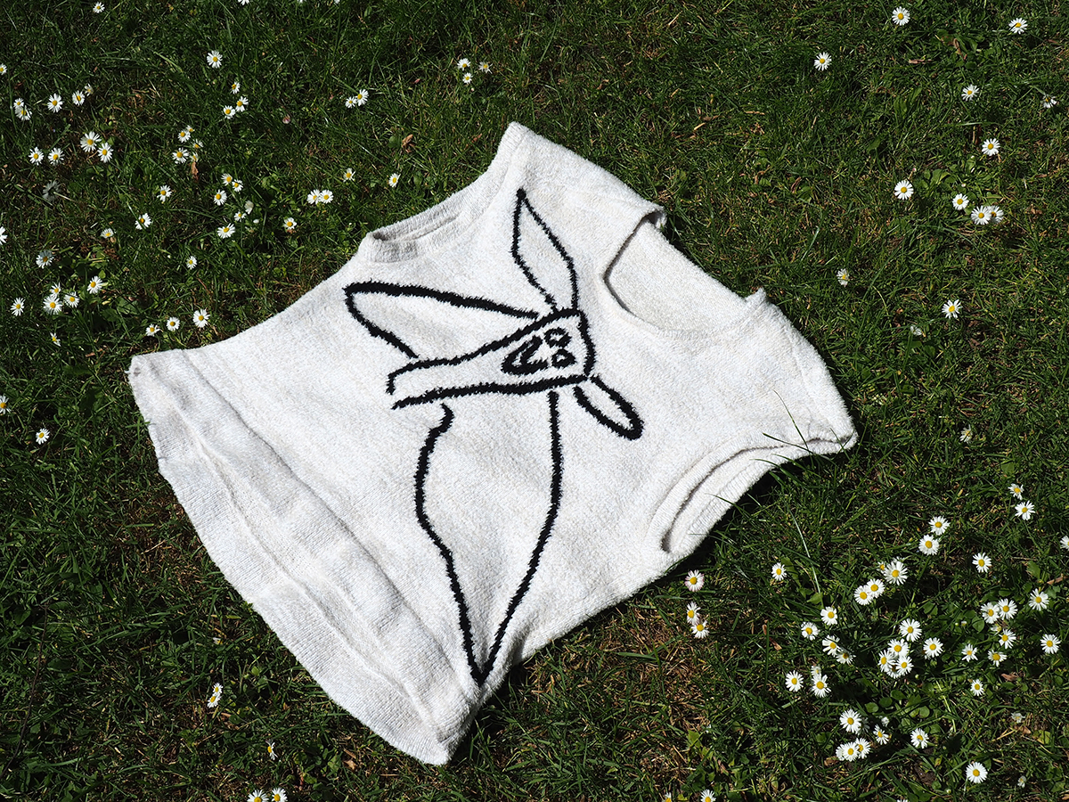 Weißer kurzärmeliger Pullover mit schwarzer, grafischer Tierzeichnung liegt auf grünem Rasen mit verstreuten weißen Gänseblümchen; Draufsicht bei natürlichem Licht.