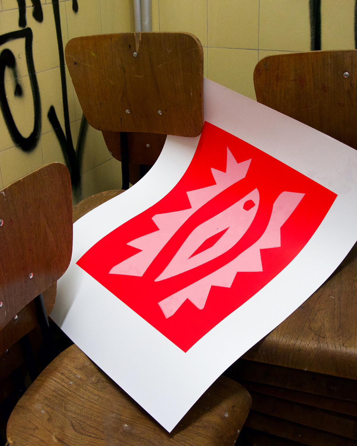 Großformatiger Druck mit rotem Rechteck und stilisiertem, hellrosa Augenmotiv lehnt auf gestapelten Holzstühlen; Hintergrund mit gelb gefliester Wand und schwarzem Graffiti.