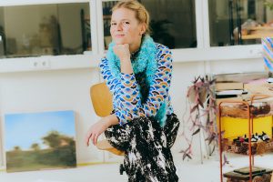 Fotografie einer Frau, die in einem hellen Atelier auf einem Stuhl sitzt. Sie trägt ein gemustertes Oberteil, einen schwarzen Rock und schwarze Stiefel; im Hintergrund stehen Regale, Pflanzen und ein Rollwagen mit Materialien.