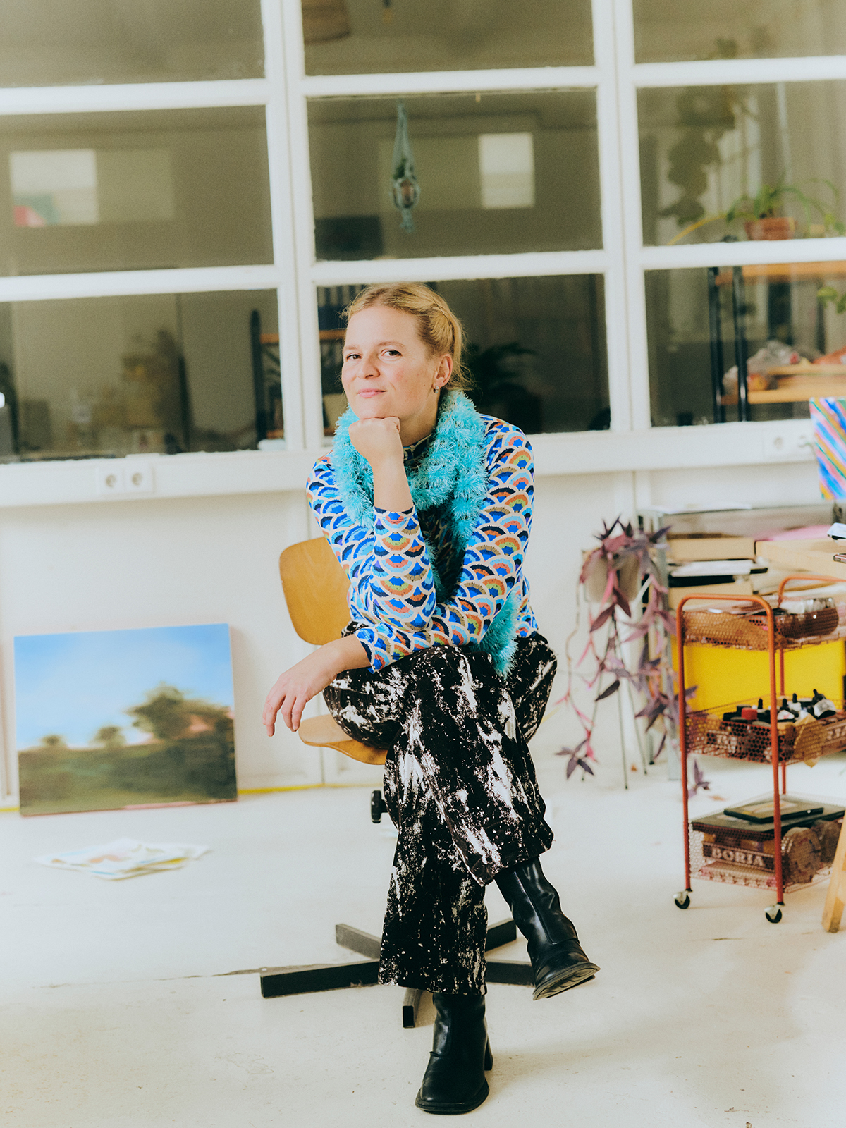 Fotografie einer Frau, die in einem hellen Atelier auf einem Stuhl sitzt. Sie trägt ein gemustertes Oberteil, einen schwarzen Rock und schwarze Stiefel; im Hintergrund stehen Regale, Pflanzen und ein Rollwagen mit Materialien.