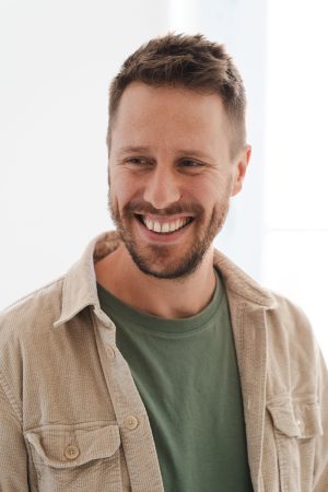 Mann mit Bart vor hellem Hintergrund, beiges Hemd mit Olivfarbenen Shirt, lockerer Stil, entspannt lächelnd