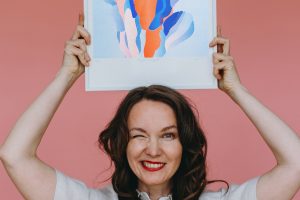 Porträt einer lächelnden Frau vor rosafarbenem Hintergrund; sie hält über ihrem Kopf einen gerahmten Kunstdruck mit abstrakten Formen in Blau-, Rosa- und Orangetönen.