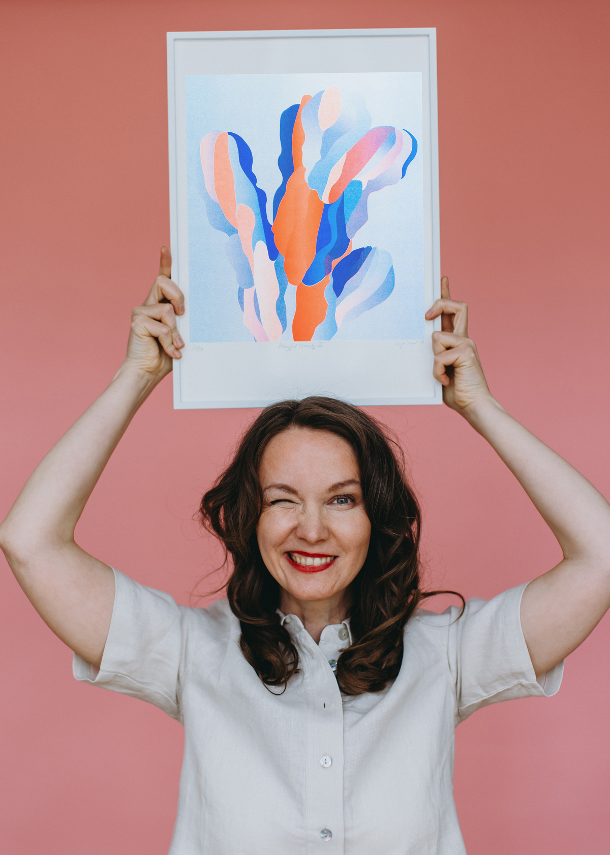 Porträt einer lächelnden Frau vor rosafarbenem Hintergrund; sie hält über ihrem Kopf einen gerahmten Kunstdruck mit abstrakten Formen in Blau-, Rosa- und Orangetönen.