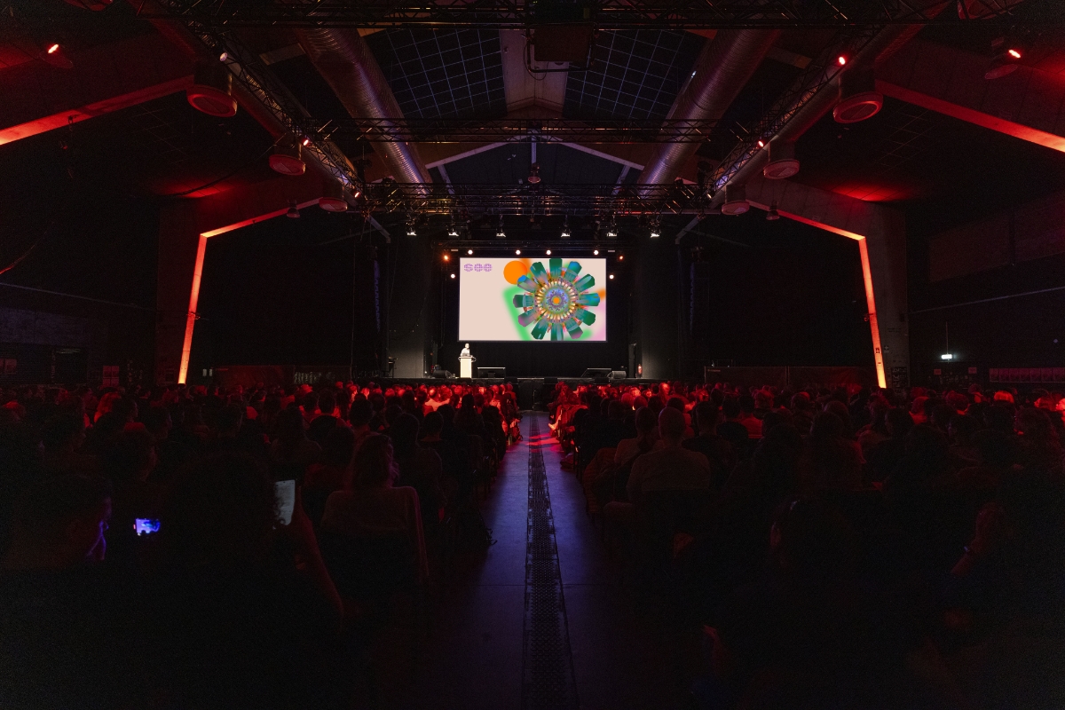 Große Konferenzhalle mit roter Bühnenbeleuchtung: Ein Speaker steht am Rednerpult vor einer großen Leinwand mit farbiger Grafik, während ein großes Publikum in Reihen davor sitzt und der Präsentation folgt.