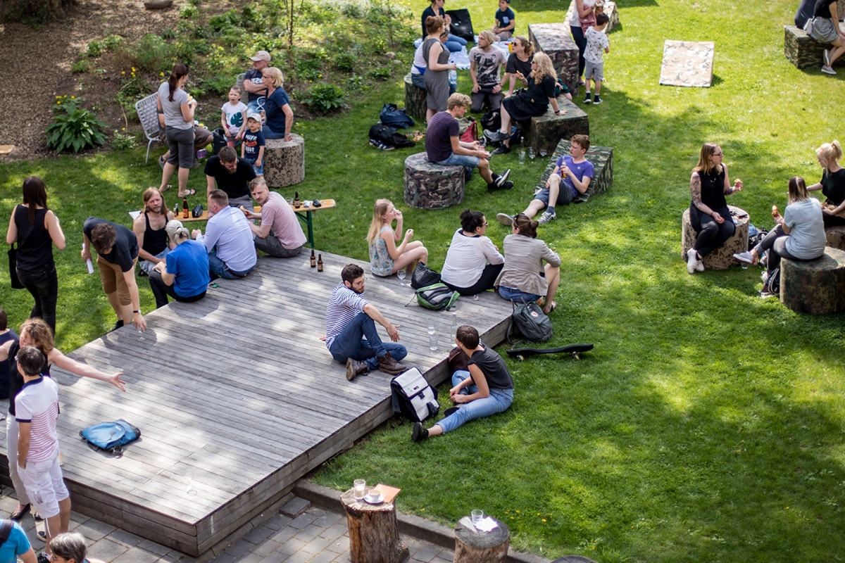 Menschen sitzen und stehen in kleinen Gruppen auf einer grünen Wiese und auf einer Holzplattform in einem Park oder Garten, unterhalten sich entspannt und genießen eine informelle Zusammenkunft im Freien.