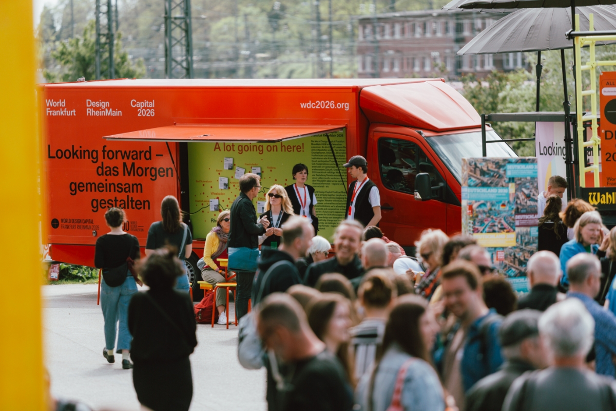 Roter Informations-Truck zur „World Design Capital Frankfurt RheinMain 2026“ steht bei einer Veranstaltung im Freien; davor informieren sich Besucherinnen und Besucher an einem geöffneten Infostand, während sich eine größere Menschenmenge auf dem Gelände bewegt.
