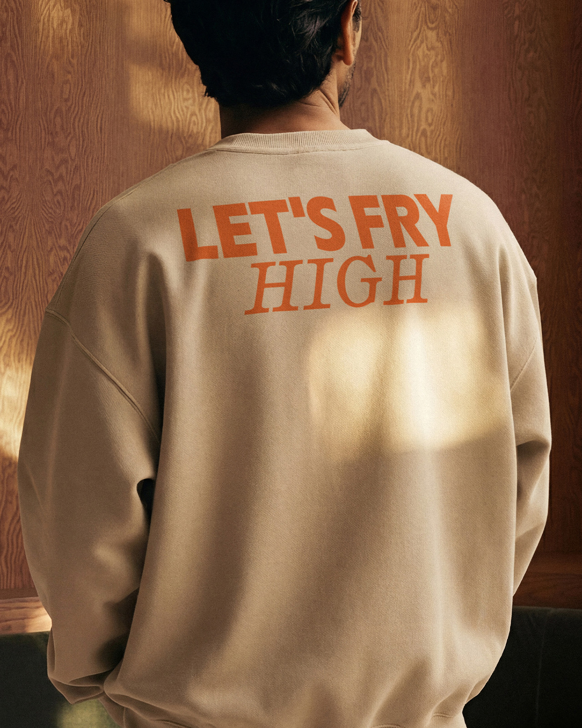 Merchandise-Sweater für The Fritten. Eine Person trägt einen beigefarbenen Oversize-Sweatshirt mit dem Rücken zur Kamera. Auf dem Rücken ist in großen orangefarbenen Buchstaben der Schriftzug »LET'S FRY HIGH« aufgedruckt, in zwei Schriften gesetzt. Der warme Hintergrund zeigt eine Holzverkleidung.