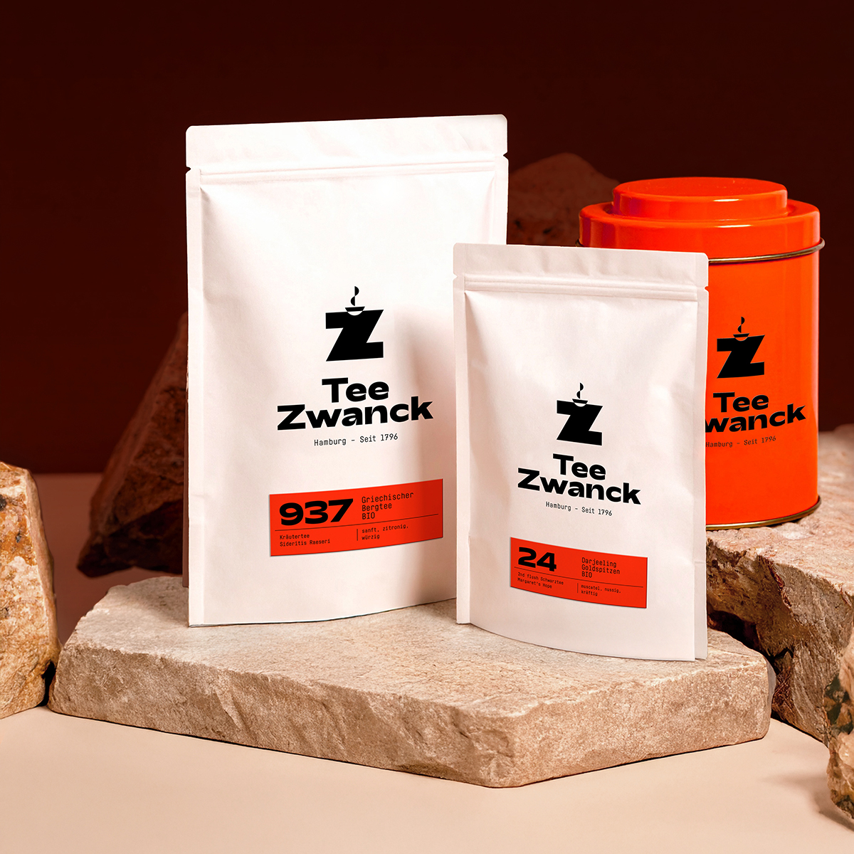 Produktsetting für Tee Zwanck. Zwei weiße Standbodenbeutel in verschiedenen Größen und eine orangefarbene Metalldose mit Z-Logo sind auf Natursteinen arrangiert. Alle Produkte zeigen das neue Markendesign mit schwarzem Z-Logo und orangefarbenen Etiketten, im Hintergrund ein warmer dunkler Hintergrund.