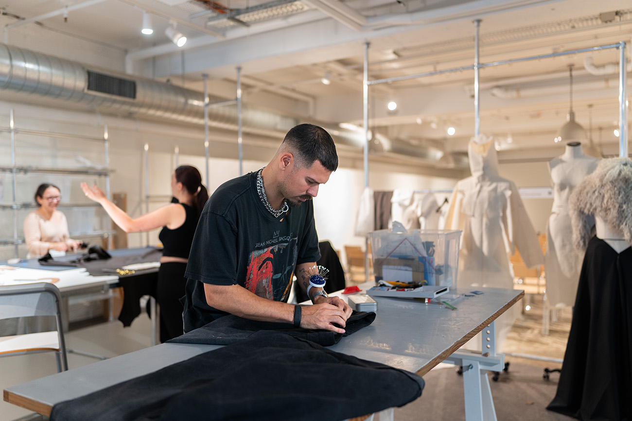 Ein junger Mann in schwarzem T-Shirt arbeitet konzentriert an einem Schneidetisch und befestigt Stecknadeln an schwarzem Stoff. Im Hintergrund arbeiten zwei weitere Personen in einem hellen Modedesign-Atelier mit Schneiderpuppen.