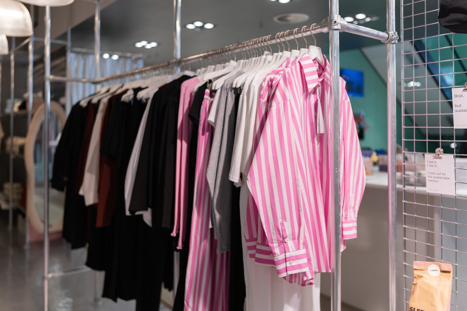Kleiderstange mit verschiedenen Kleidungsstücken, darunter auffällige pink-weiß gestreifte Hemden und dunkle Basics. Im Hintergrund ist ein Schild mit der Aufschrift „I see it. I like it. I look out for the sustainable version." zu sehen.