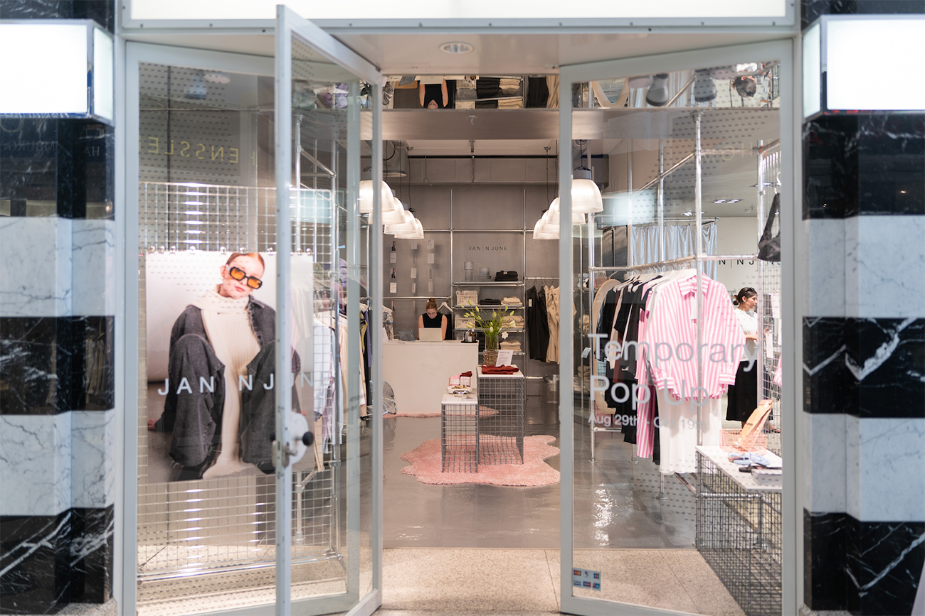 Blick durch eine gläserne Drehtür in einen hellen Pop-up-Store von Jan 'N June. Die Schaufensterscheibe trägt den Hinweis „Temporary Pop-up, Aug 29th – Oct 19th". Im Inneren sind Kleiderstangen mit pink-weißen Hemden und eine Verkäuferin am Tresen zu sehen.