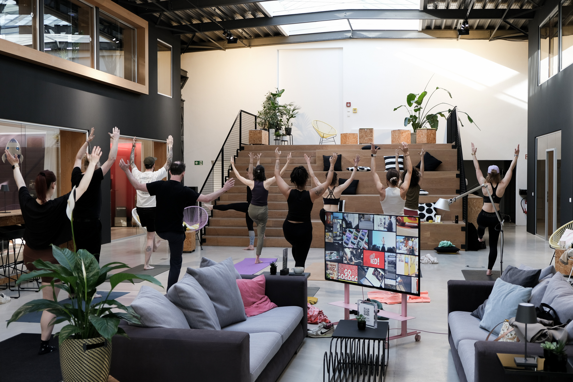 Eine Gruppe von Menschen steht in einem offenen, modernen Innenraum mit hohen Decken, Oberlichtern und Holzstufen und führt gemeinsam eine Yoga- oder Stretching-Übung aus. Die Teilnehmenden balancieren auf einem Bein und strecken die Arme nach oben. Im Vordergrund stehen Sofas und ein Bildschirm mit einer Kachelübersicht, während Pflanzen und Sitzbereiche die kreative, entspannte Atmosphäre unterstreichen.