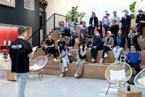 Eine Person steht im Vordergrund und spricht vor einer Gruppe, die auf gestuften Holzstufen sitzt, in einem offenen, modernen Raum mit Pflanzen und viel Tageslicht. Die Zuhörenden blicken aufmerksam nach vorn, einige halten Getränke in der Hand. Der Sprecher trägt ein schwarzes T-Shirt mit der Aufschrift „PAGE – Design. Business. Community.“; die Atmosphäre wirkt konzentriert, zugleich locker und gemeinschaftlich.