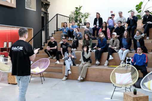 Eine Person steht im Vordergrund und spricht vor einer Gruppe, die auf gestuften Holzstufen sitzt, in einem offenen, modernen Raum mit Pflanzen und viel Tageslicht. Die Zuhörenden blicken aufmerksam nach vorn, einige halten Getränke in der Hand. Der Sprecher trägt ein schwarzes T-Shirt mit der Aufschrift „PAGE – Design. Business. Community.“; die Atmosphäre wirkt konzentriert, zugleich locker und gemeinschaftlich.