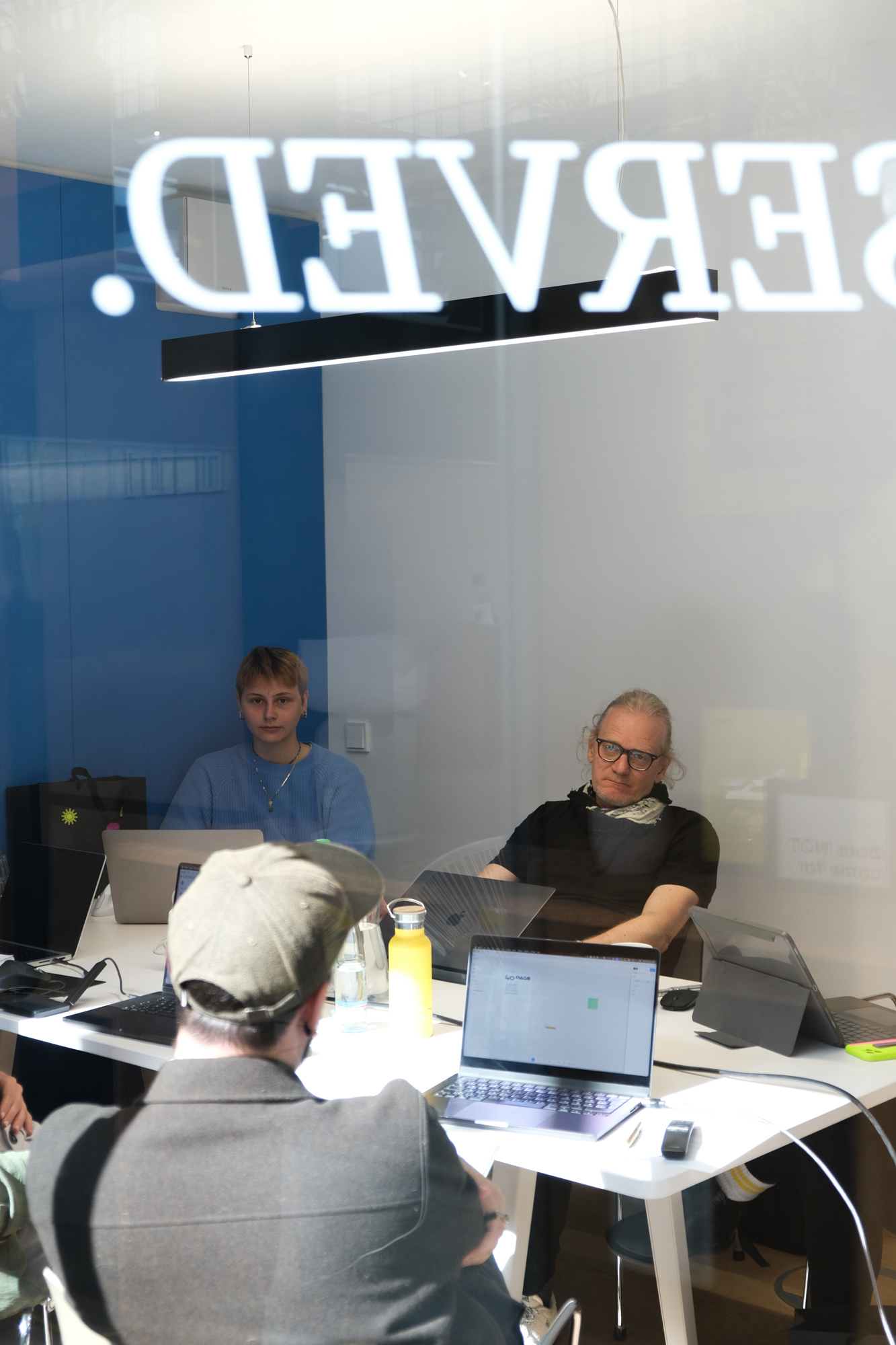 Blick durch eine Glasscheibe in einen Besprechungsraum: Drei Personen sitzen an einem Tisch und arbeiten an Laptops, während im Vordergrund eine weitere Person mit dem Rücken zur Kamera sitzt. Die spiegelnde Scheibe zeigt spiegelverkehrte Schrift und Lichtreflexe. Auf dem Tisch stehen Wasserflaschen und Arbeitsmaterialien; die Stimmung wirkt ruhig und konzentriert.