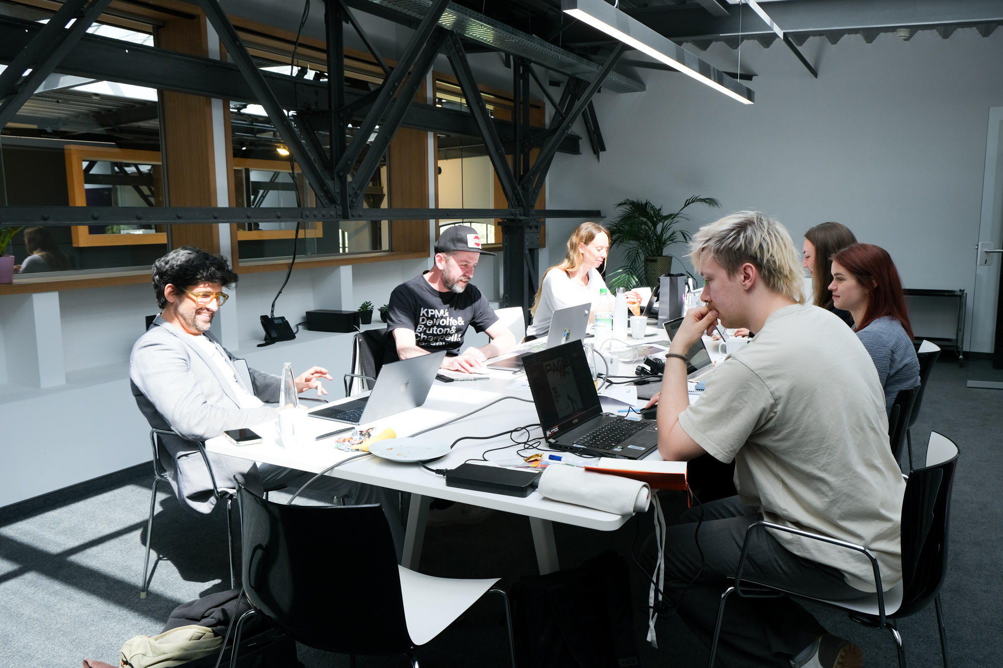 Sechs Personen sitzen an einem Tisch in einem hellen, modernen Arbeitsraum mit sichtbaren Stahlträgern und großen Fenstern und arbeiten konzentriert an Laptops. Auf dem Tisch liegen Notizbücher, Kabel und Getränke; Sonnenlicht fällt durch die Fenster und schafft eine fokussierte, produktive Atmosphäre.