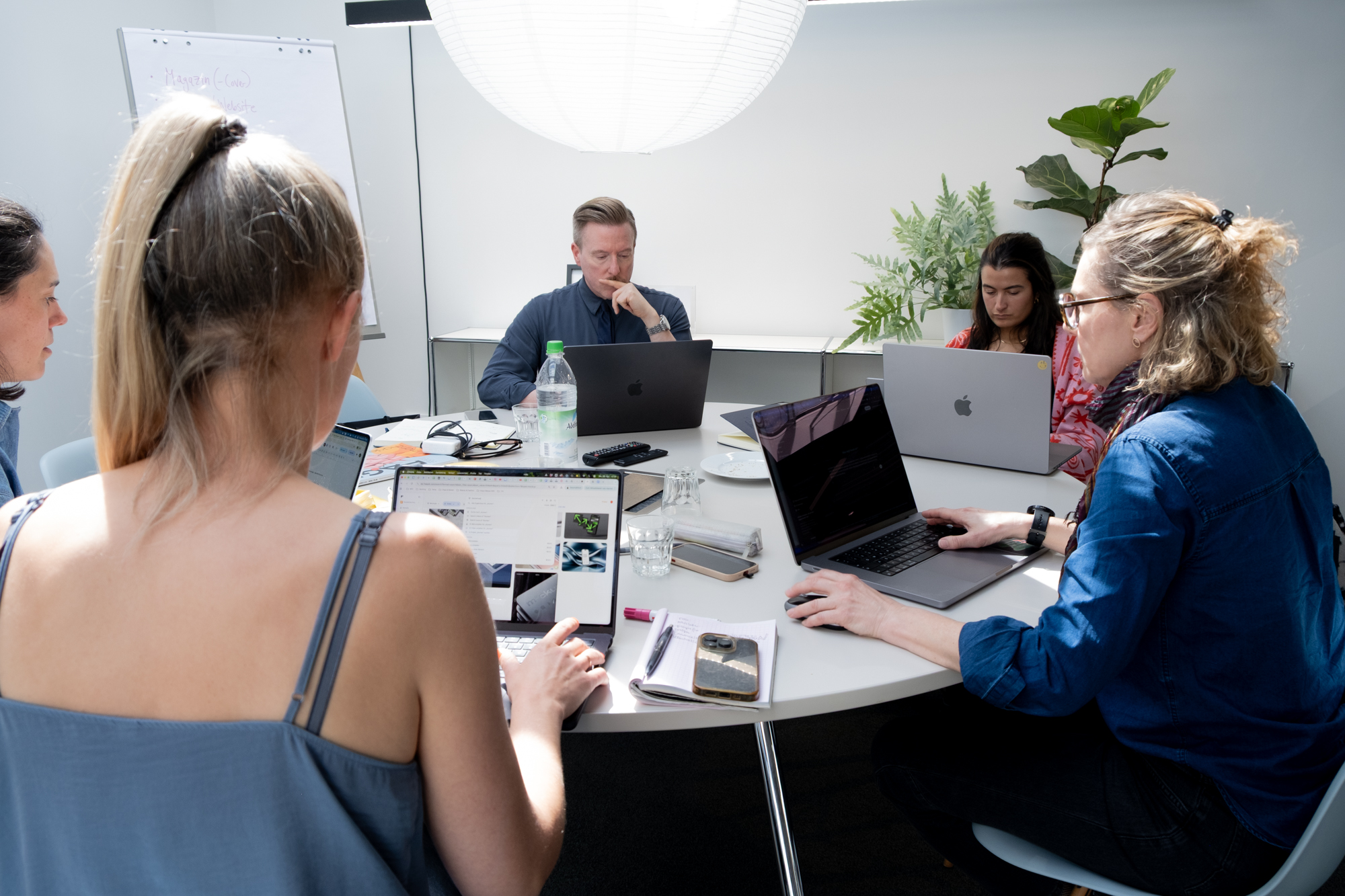 Fünf Personen sitzen an einem runden Tisch in einem hellen, minimalistischen Raum und arbeiten konzentriert an ihren Laptops. Auf dem Tisch liegen Notizbücher, Smartphones, Wasserflaschen und Gläser. Eine große runde Deckenlampe, Pflanzen und ein Whiteboard im Hintergrund schaffen eine ruhige, fokussierte Arbeitsatmosphäre.