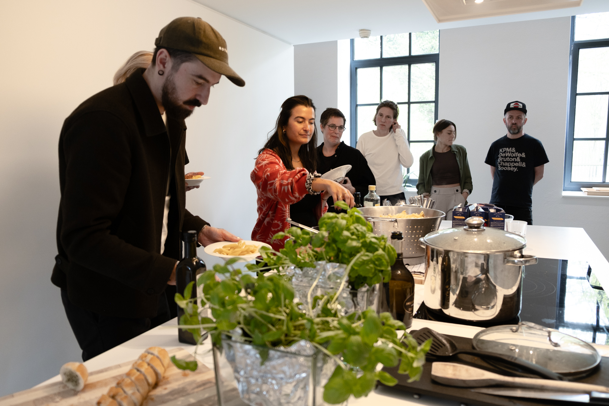 Mehrere Personen stehen in einer hellen Küche um eine Kochinsel und bereiten gemeinsam Essen zu. Im Vordergrund richtet eine Person Pasta auf einem Teller an, während eine andere Nudeln aus einem großen Topf nimmt. Auf der Arbeitsfläche liegen frische Kräuter, Brot und Kochutensilien; im Hintergrund beobachten weitere Teilnehmende das Geschehen. Die Atmosphäre wirkt gemeinschaftlich und entspannt.