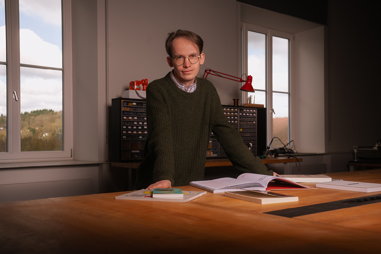 Porträt eines Mannes mit Brille, der in einem Atelier oder Arbeitsraum an einem Holztisch lehnt; vor ihm liegen mehrere Bücher und ein aufgeschlagenes Magazin, im Hintergrund sind Regale mit Materialien und eine rote Schreibtischlampe zu sehen, während durch die Fenster Tageslicht einfällt.