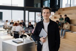 Porträt eines jungen Mannes in einem modernen Büro mit offenen Sitzstufen; er hält einen Laptop in der Hand, während im Hintergrund mehrere Menschen an Tischen zusammenarbeiten.