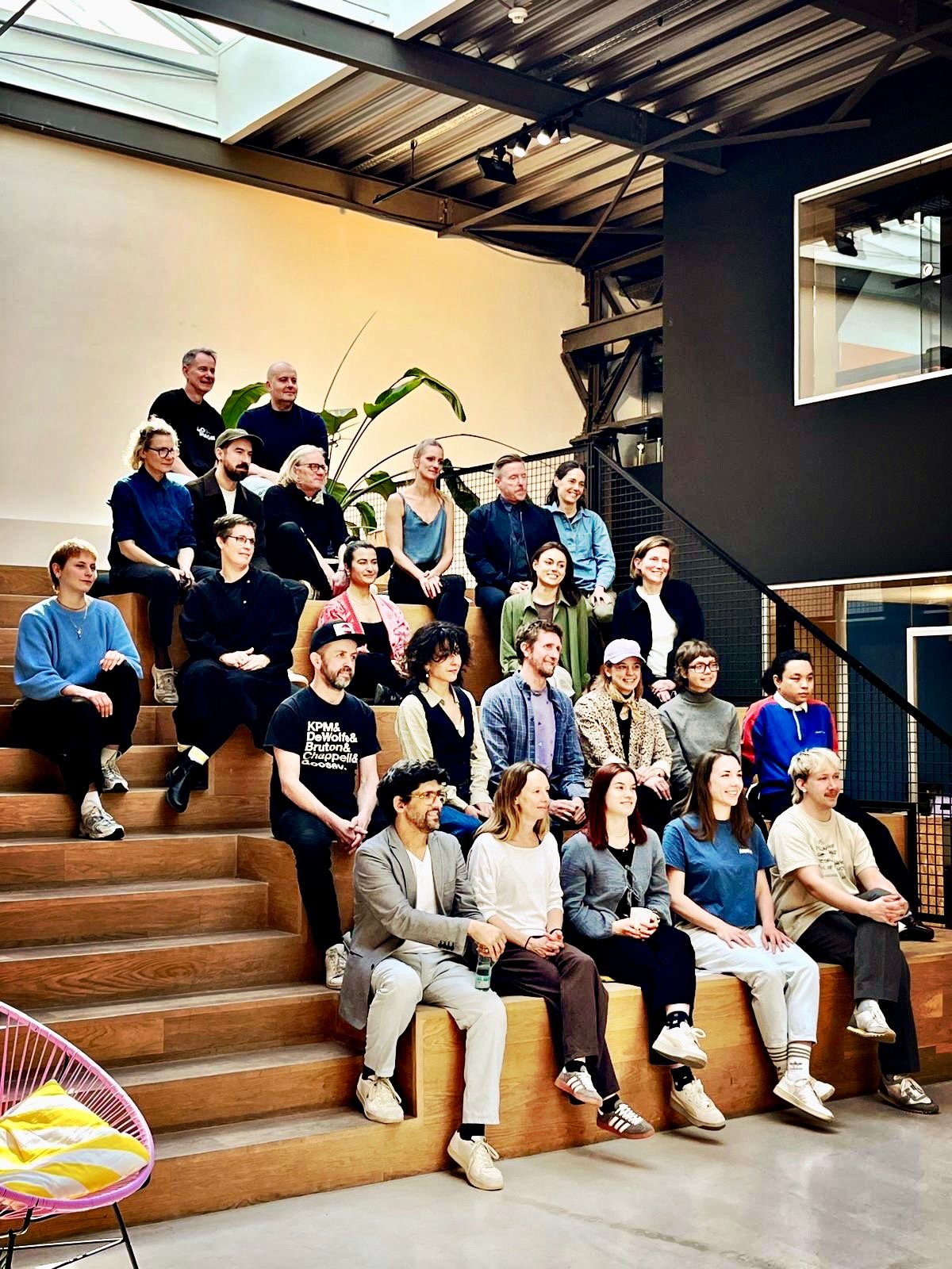 Eine größere Gruppe von Menschen sitzt gestaffelt auf breiten Holzstufen in einem modernen Innenraum mit hohen Decken, Stahlträgern und Oberlichtern. Die Teilnehmenden blicken nach vorn, einige lächeln, als würden sie für ein Gruppenfoto posieren. Pflanzen und eine minimalistische Architektur prägen die Umgebung; die Atmosphäre wirkt offen, kreativ und gemeinschaftlich.