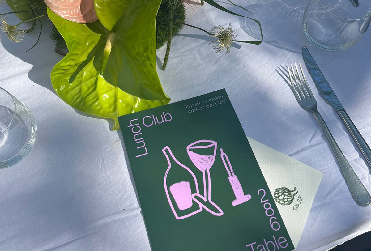 Das dunkelgrüne Menüheft des Lunch Club Table in Amsterdam West liegt auf einem weiß gedeckten Tisch, daneben Besteck und eine grüne Blumenvase. Das Heft zeigt die pinken Linienillustrationen von Flasche, Weinglas und Kerze sowie den Schriftzug »Lunch Club Table 286« und den Hinweis »Private Location Amsterdam West«.