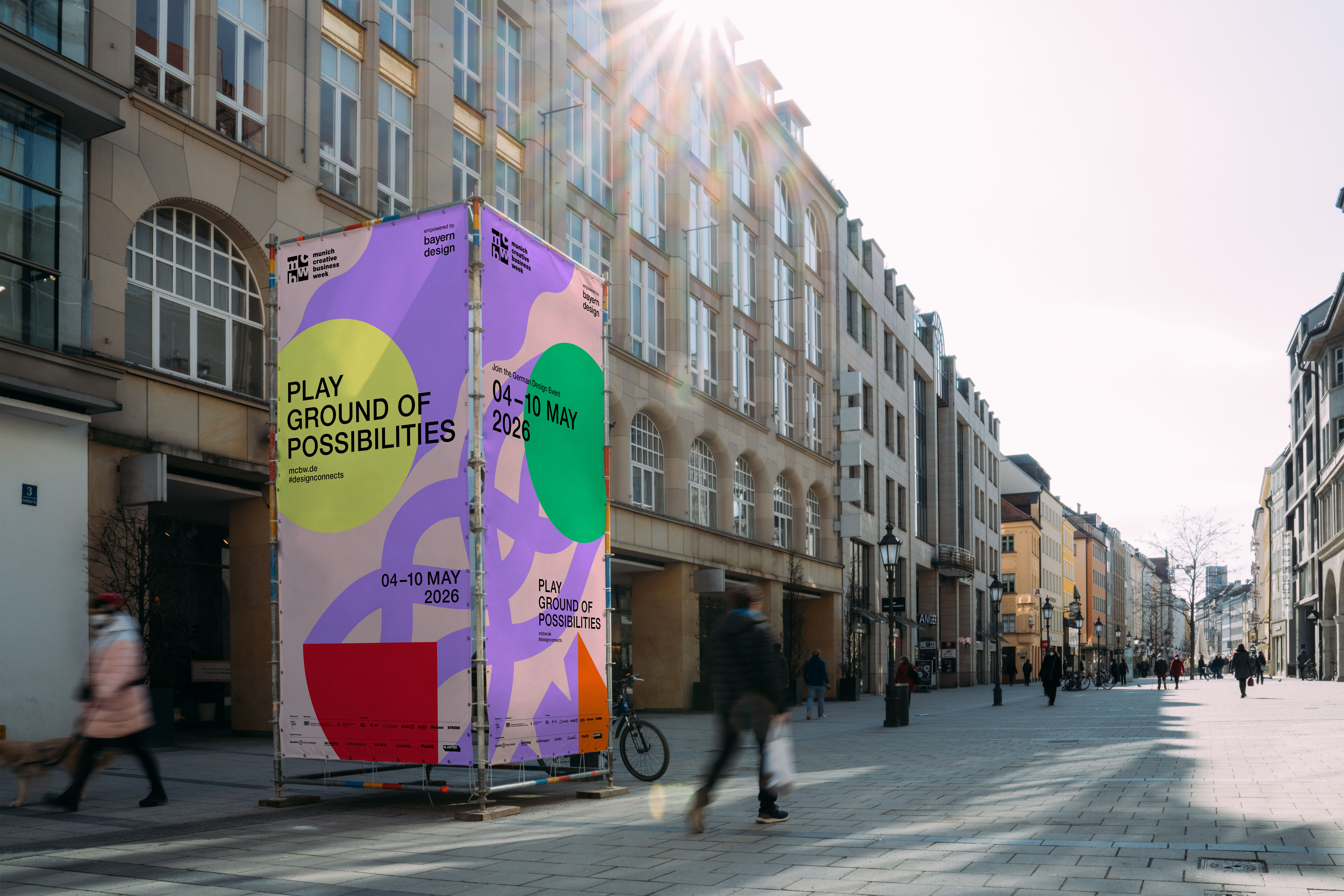 Großformatiges, farbenfrohes Werbeplakat der Munich Creative Business Week steht als Installation auf einer breiten Münchner Straße zwischen historischen Gebäuden. Das Plakat zeigt abstrakte Formen in Lila, Grün, Gelb und Rot sowie den Schriftzug „Playground of Possibilities“ und das Datum „04–10 May 2026“. Sonnenstrahlen fallen von oben ins Bild, während Passant:innen vorbeigehen und eine urbane, lebendige Atmosphäre entsteht.