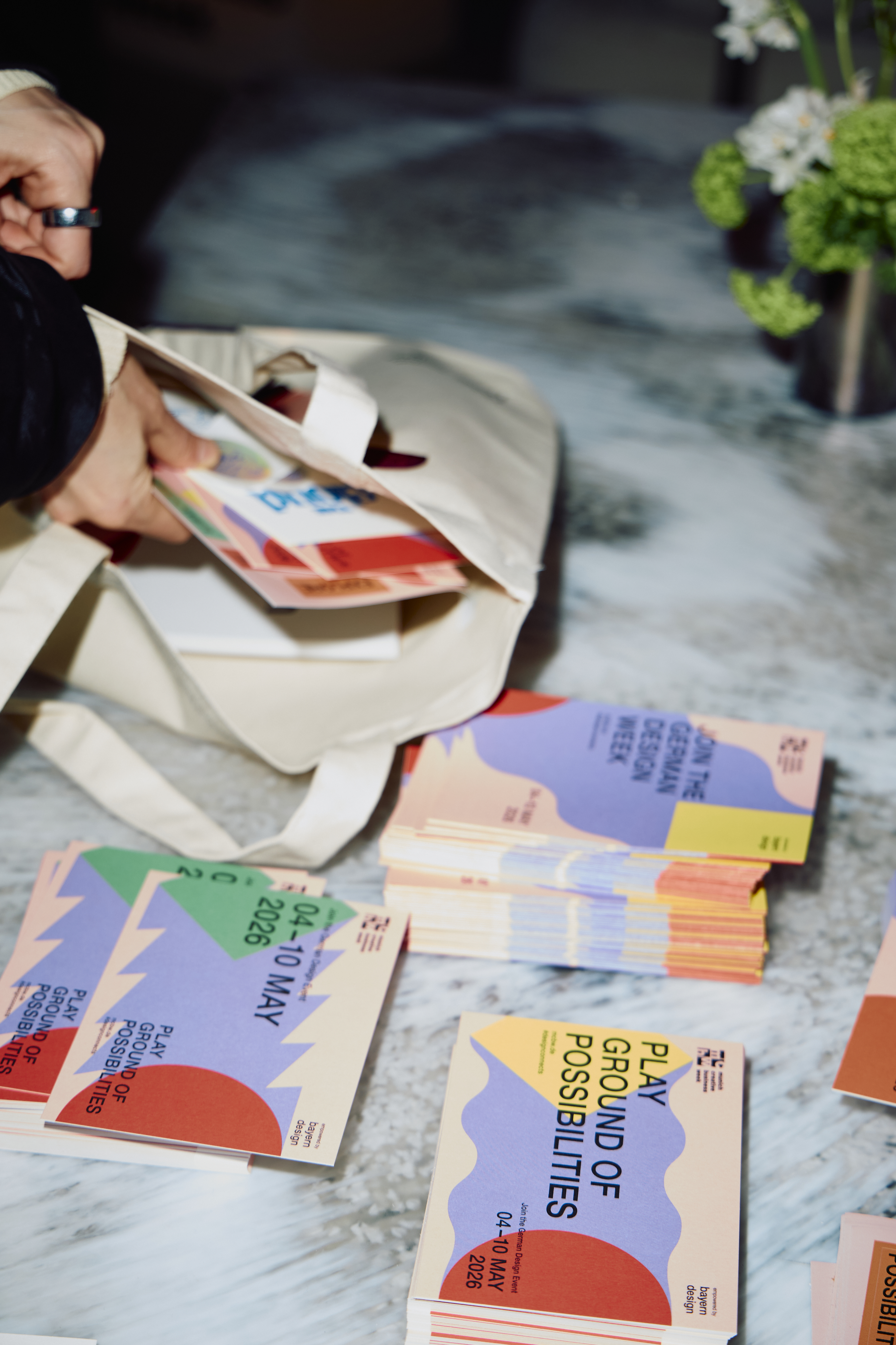 Mehrere Stapel farbenfroher Flyer und Postkarten der Munich Creative Business Week liegen auf einem Marmortisch. Eine Person greift in eine Stofftasche und legt weiteres Material dazu. Die Drucksachen zeigen abstrakte Formen und den Schriftzug „Playground of Possibilities“ sowie das Datum „04–10 May 2026“. Im Hintergrund steht eine kleine Vase mit Blumen.