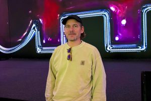 Ein Mann steht vor einer großen, leuchtenden Medieninstallation mit neonartigen Linien in Blau, Pink und Violett. Er trägt ein helles Sweatshirt, eine schwarze Kappe und hat eine Sonnenbrille am Kragen hängen. Die farbintensive, abstrakte Hintergrundgrafik erinnert an digitale Kunst und taucht die Szene in ein futuristisches Licht.