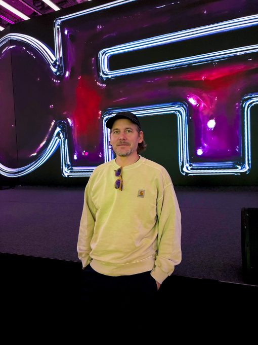 Ein Mann steht vor einer großen, leuchtenden Medieninstallation mit neonartigen Linien in Blau, Pink und Violett. Er trägt ein helles Sweatshirt, eine schwarze Kappe und hat eine Sonnenbrille am Kragen hängen. Die farbintensive, abstrakte Hintergrundgrafik erinnert an digitale Kunst und taucht die Szene in ein futuristisches Licht.