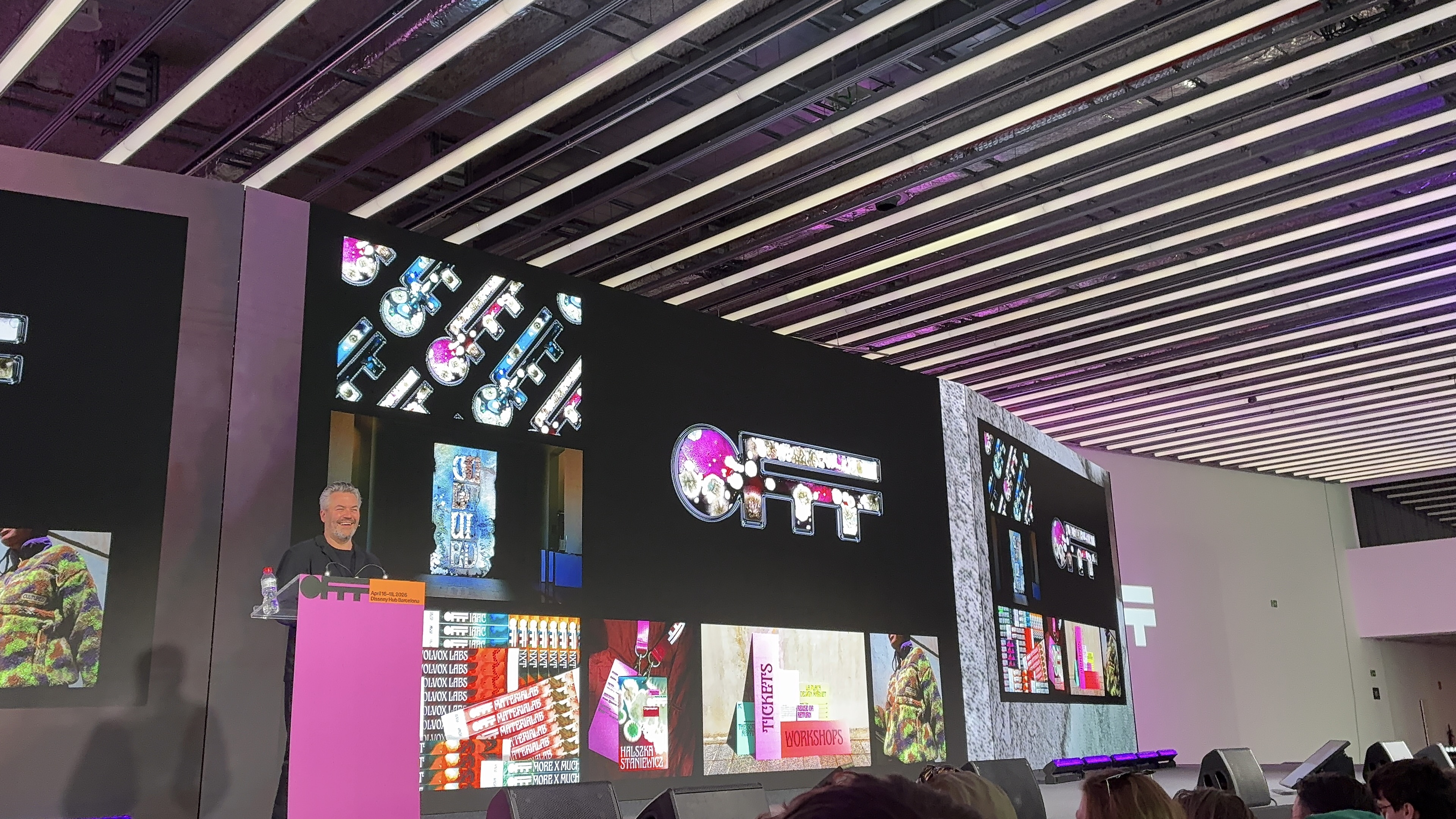 Ein Referent steht auf einer Bühne hinter einem Pult und präsentiert vor einer großen LED-Wand. Die Bildschirme zeigen farbenfrohe, grafische Arbeiten und Typografie, darunter Schlüssel-Illustrationen und Layouts. Im Vordergrund sitzt Publikum, während die moderne Halle mit langen Lichtleisten an der Decke eine klare, technische Atmosphäre schafft.