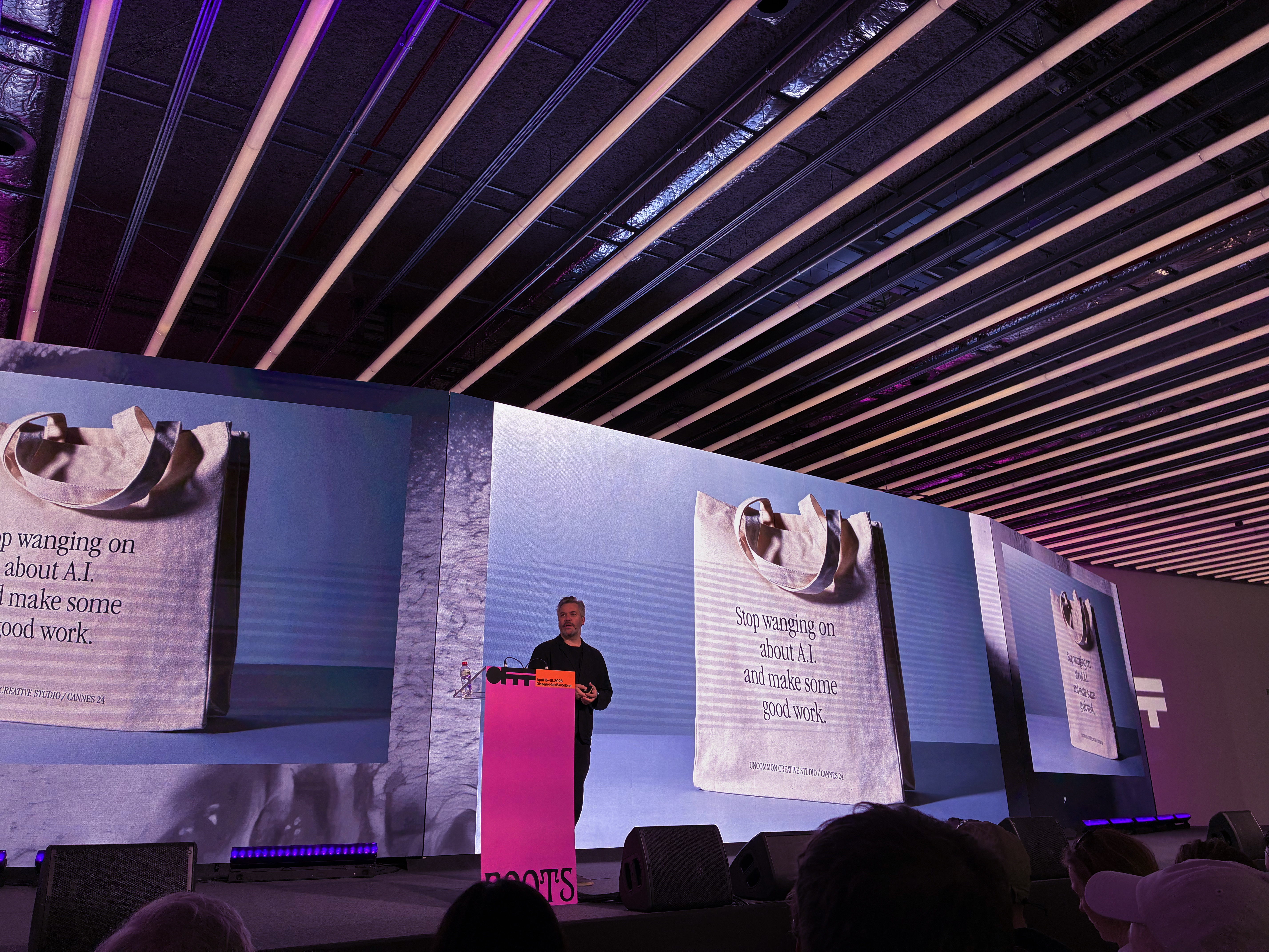 Ein Referent steht auf einer Bühne hinter einem pinken Rednerpult und präsentiert vor mehreren großen Bildschirmen. Auf den Displays ist eine Stofftasche mit der Aufschrift „Stop wanging on about A.I. and make some good work.“ zu sehen. Die moderne Veranstaltungshalle ist mit langen Lichtleisten an der Decke ausgestattet, im Vordergrund sitzt das Publikum und verfolgt den Vortrag.