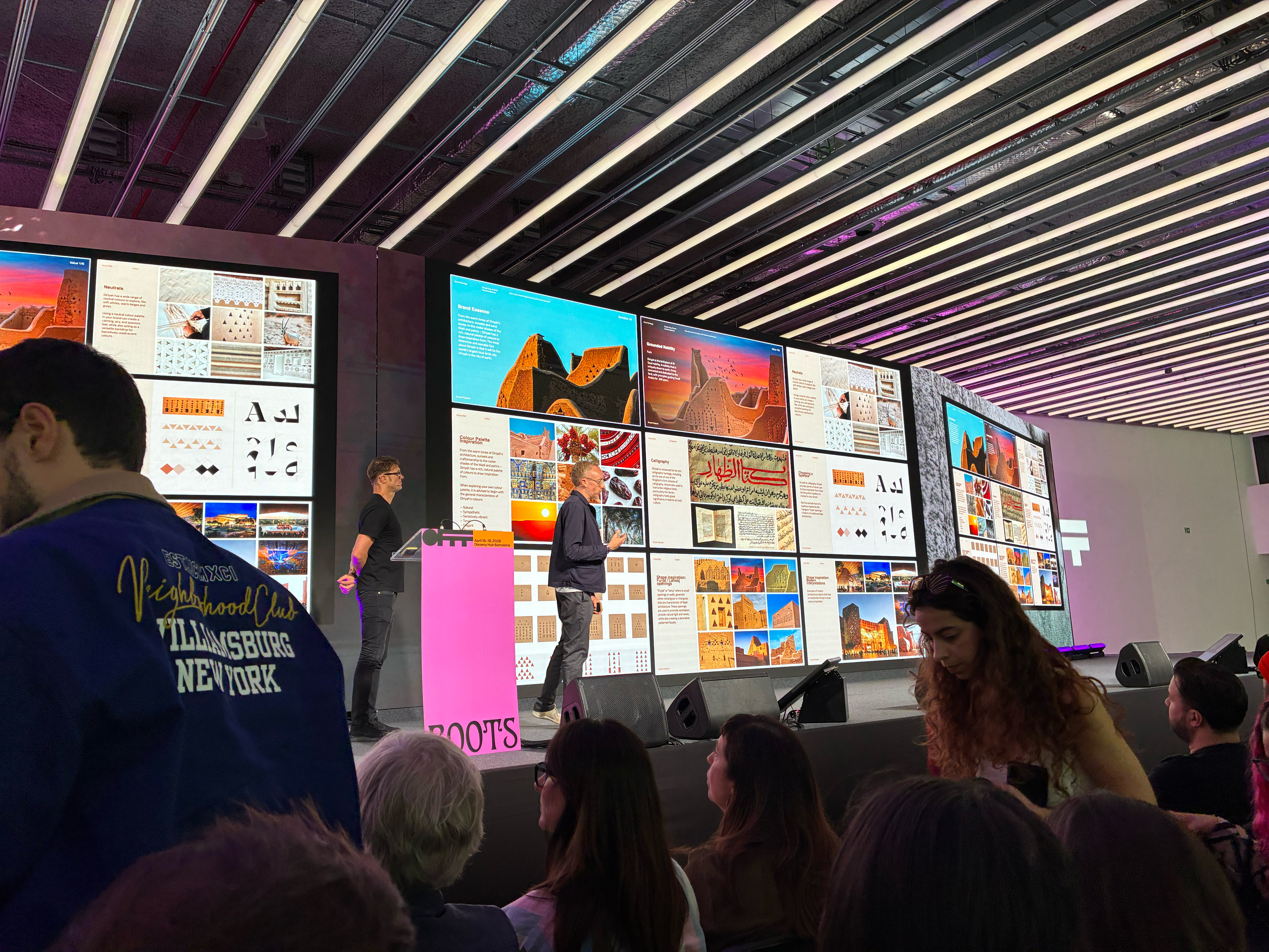 Zwei Referenten stehen auf einer Bühne und präsentieren vor einer großen, mehrteiligen LED-Wand mit zahlreichen Bildern, Grafiken und Designbeispielen. Die Screens zeigen Collagen aus Architektur, Typografie und visuellen Elementen in kräftigen Farben. Im Vordergrund sitzt das Publikum und verfolgt den Vortrag, während die moderne Halle mit langen Lichtleisten an der Decke beleuchtet ist.