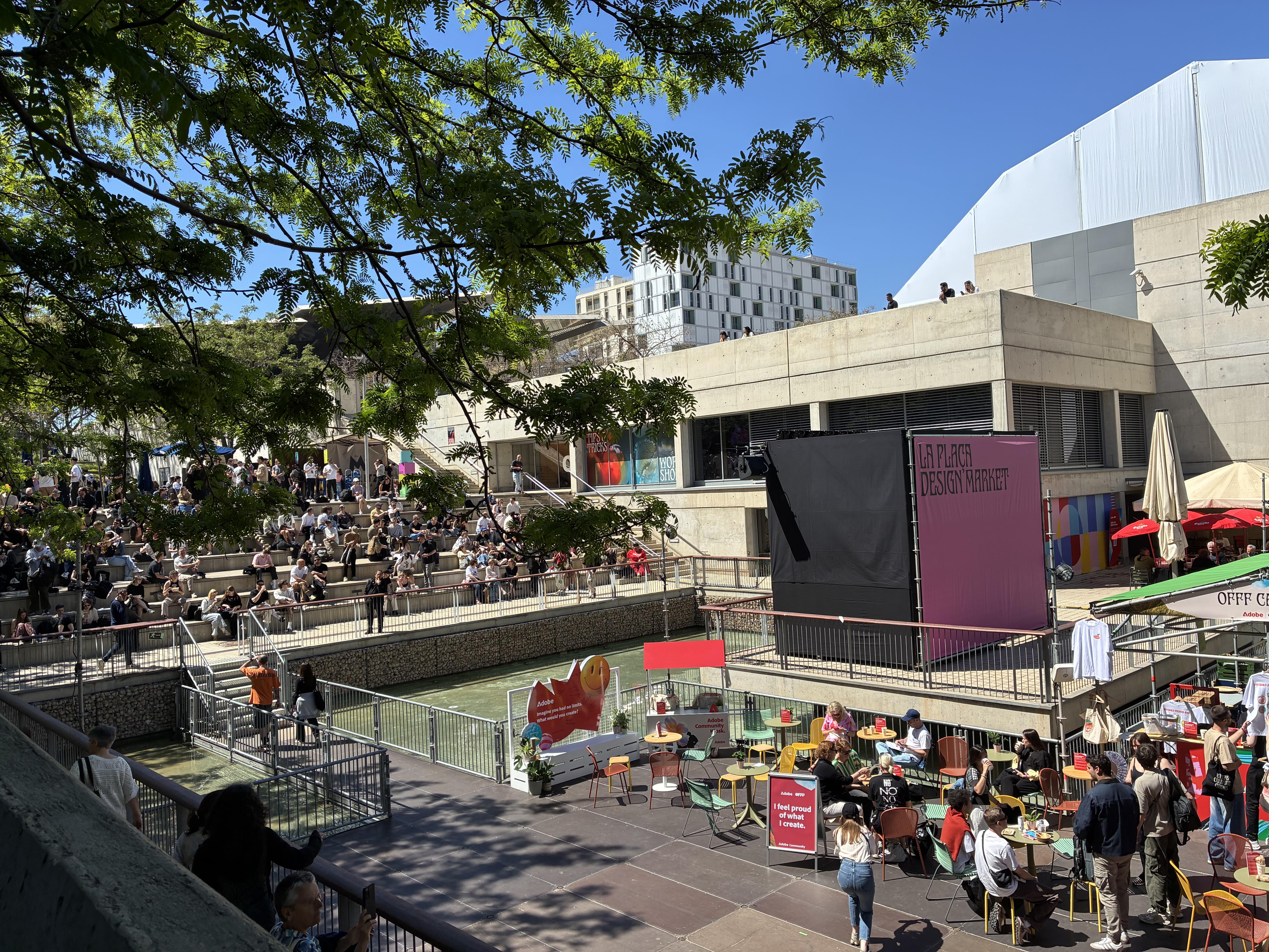 Belebter Außenbereich eines Design-Events mit vielen Menschen auf Treppenstufen und entlang eines Wasserbeckens. Besucher:innen sitzen, stehen oder gehen über mehrere Ebenen eines modernen Gebäudekomplexes. Im Vordergrund gibt es eine kleine Café- und Marktfläche mit Tischen, Stühlen und Ständen, darunter ein Bereich mit der Beschriftung „La Plaza Design Market“. Bäume spenden Schatten, die Szene wirkt sonnig, offen und lebendig.