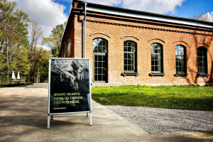 Ein freistehendes Werbeschild steht auf einem gepflasterten Weg vor einem eingeschossigen Backsteingebäude mit hohen, rundbogenförmigen Fenstern. Auf dem Schild ist ein grafisches Motiv mit der Aufschrift „BRAVE HEARTS DARE TO DESIGN. YOU ARE HERE.“ sowie „ISARWINKEL“ zu sehen. Das Gebäude liegt in einer grünen Umgebung mit Bäumen und Rasenflächen, darüber ein blauer Himmel mit weißen Wolken.