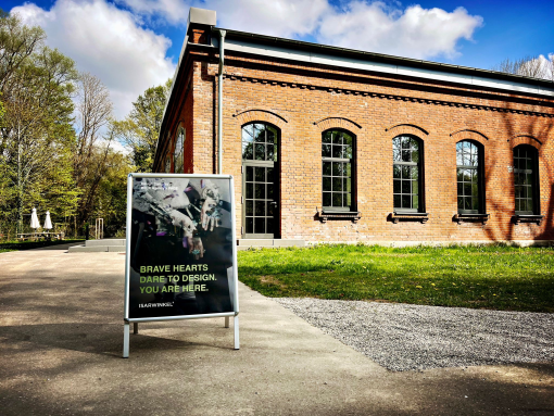 Ein freistehendes Werbeschild steht auf einem gepflasterten Weg vor einem eingeschossigen Backsteingebäude mit hohen, rundbogenförmigen Fenstern. Auf dem Schild ist ein grafisches Motiv mit der Aufschrift „BRAVE HEARTS DARE TO DESIGN. YOU ARE HERE.“ sowie „ISARWINKEL“ zu sehen. Das Gebäude liegt in einer grünen Umgebung mit Bäumen und Rasenflächen, darüber ein blauer Himmel mit weißen Wolken.
