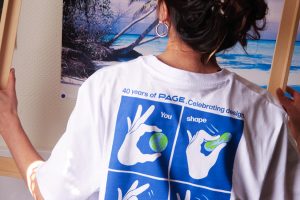 Person von hinten vor einem tropischen Strandposter, trägt ein weißes T-Shirt mit blauem, vierteiligem Comic-Motiv: Hände formen eine Erde und den Schriftzug „You shape the world. Do it with love!“ sowie „40 years of PAGE. Celebrating design.“