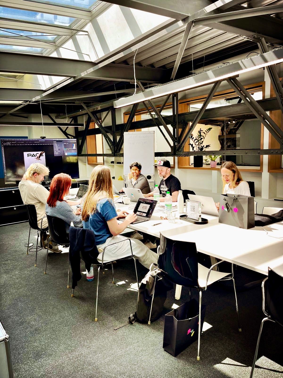 Sechs Personen sitzen an einem langen Tisch in einem hellen, modernen Workshopraum mit Dachfenstern und sichtbaren Stahlträgern. Sie arbeiten konzentriert an Laptops und Notizen, während auf einem großen Bildschirm im Hintergrund eine Präsentation mit dem PAGE-Logo zu sehen ist. Auf dem Tisch stehen Wasserflaschen, Taschen und Arbeitsmaterialien; die Atmosphäre wirkt kollaborativ und kreativ.