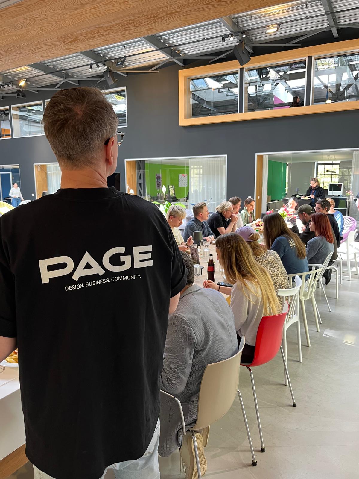 Eine Person steht im Vordergrund mit dem Rücken zur Kamera und trägt ein schwarzes T-Shirt mit der Aufschrift „PAGE – Design. Business. Community.“. Vor ihr sitzt eine größere Gruppe von Menschen an einem langen Tisch in einem hellen, modernen Raum mit großen Fenstern und Galerieebene. Die Teilnehmenden sind im Gespräch, essen und arbeiten gemeinsam; die Atmosphäre wirkt offen, kommunikativ und gemeinschaftlich.