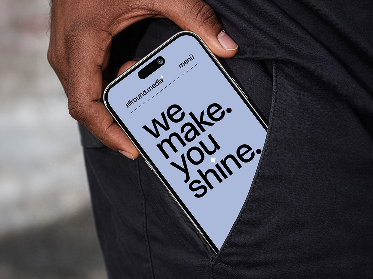Website-Mockup der Agentur allround.media, dargestellt auf einem Smartphone, das eine Hand halb in eine Hosentasche schiebt. Der Bildschirm zeigt auf hellblauem Hintergrund den Claim »we make. you shine.« in großer, fetter schwarzer Schrift sowie die Navigation mit Logo und Menü-Button.