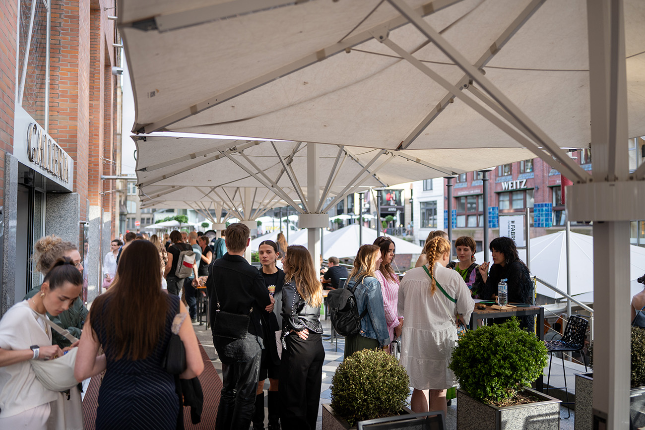 Belebte Außenterrasse eines Restaurants in der Hamburger Innenstadt, umgeben von Backsteingebäuden. Viele modisch gekleidete Menschen stehen und sitzen unter großen weißen Sonnenschirmen; im Hintergrund ist das Schild „Weitz" zu sehen.