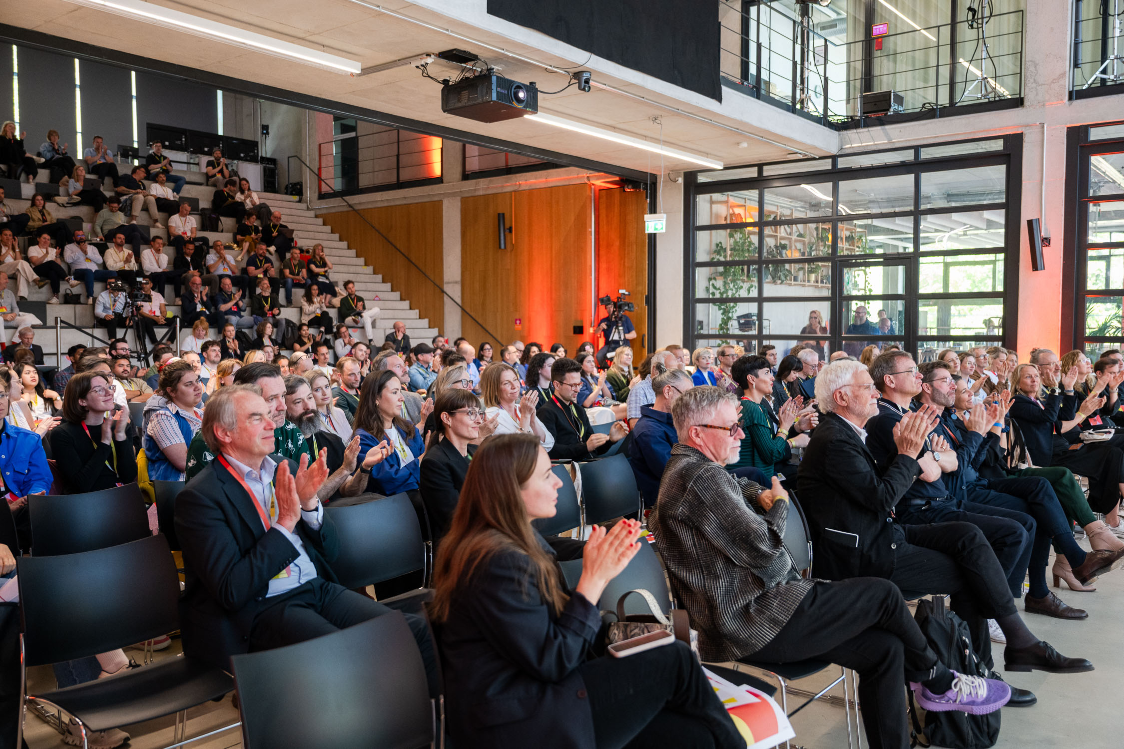 Großer, moderner Veranstaltungsraum mit ansteigenden Sitzreihen und vielen Besucher:innen, die applaudieren. Das Publikum sitzt dicht beieinander, viele tragen Namensbadges und blicken nach vorne zur Bühne (nicht im Bild). Im Hintergrund sind Kameras, Glaswände und eine offene Galerieebene zu sehen, die dem Raum eine industrielle, lichtdurchflutete Atmosphäre geben.
